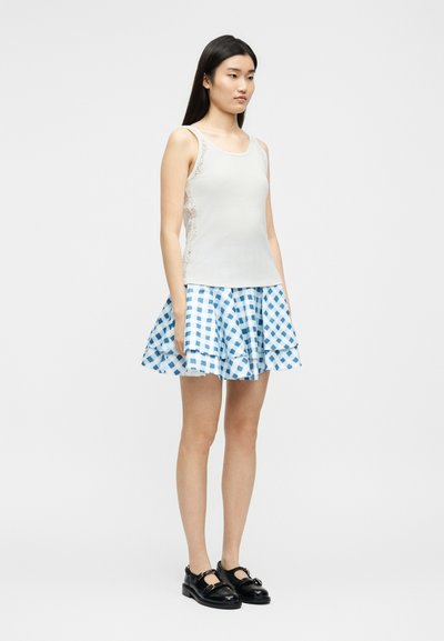 Γυναίκα που φοράει λευκό αμάνικο τοπ με δαντέλα στα πλάγια, μπλε και λευκή καρό φούστα με καμπάνα και μαύρα παπούτσια Mary Jane, στέκεται μπροστά σε απλό φόντο.