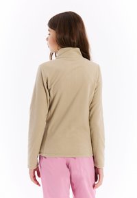 Beige fleece jack met een staande kraag en lange mouwen, met een zachte textuur. Draagt roze broek, gezien van achteren.