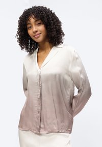 Γυναίκα με σγουρά μαλλιά, που φοράει μπλούζα με μακριά μανίκια σε αποχρώσεις από μπεζ έως ανοιχτό καφέ, στέκεται με τα χέρια πίσω από την πλάτη της μπροστά σε απλό φόντο.