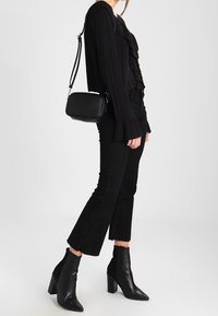 Blouse noire à volants, jean noir taille haute et court, et bottines noires à talons blocs. Petit sac bandoulière noir avec sangle.