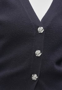 Dark navy cardigan with three silver hexagonal buttons featuring a raised floral design fastened over a white top.