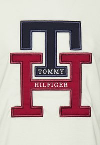 Biały sweatshirt z dużym haftowanym logo z granatowymi i czerwonymi literami "T" i "H," umieszczonymi nad napisem "TOMMY HILFIGER" w białym kolorze.