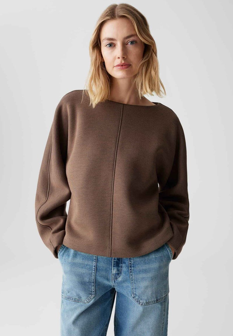 Maglione marrone con scollatura ampia, spalle scese e finitura testurizzata, abbinato a jeans di denim blu chiaro con tasche a toppa.