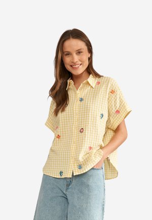 Naeratav naine, kes kannab lahtist kollast ruudulist lühikese varrukaga särki, millel on värviliste mereloomade mustrid, ning helesiniseid teksapükse, käsi taskus.