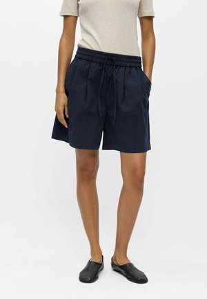 Personne portant un haut beige à manches courtes, un short bleu marine avec cordon et poches, et des chaussures noires sans lacets, debout devant un fond uni.