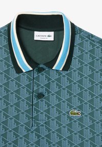 Grön mönstrad polo-shirt med ljusblå och krämfärgade randiga krage. Har två svarta knappar och en liten broderad krokodil-logotyp.