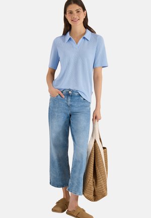 Kvinde iført lyseblå kortærmet skjorte med krave, brede cropped jeans, beige slip-on sko, holder en stor flettet tote-taske.