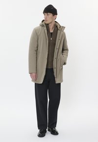 Beige isolerad kappa med huva, dragkedja och tryckknappar, bärs över en brun zip-up tröja, tillsammans med svarta byxor och stövlar.