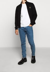 Svart zip-up jacka med hög krage, vit t-shirt under, blå jeans och svarta läderankelstövlar.