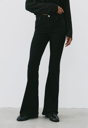 Personne portant un jean noir taille haute évasé avec des chaussures noires et un haut noir à manches longues, debout devant un fond clair uni.