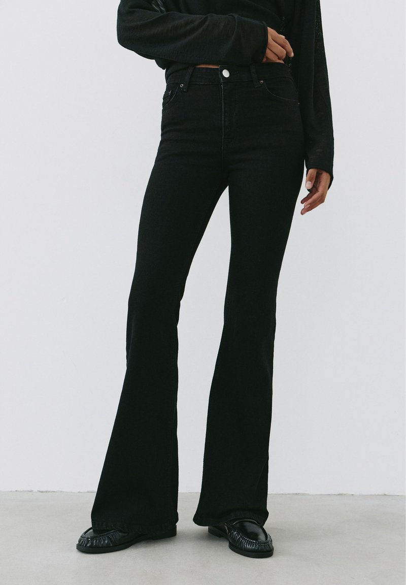Personne portant un jean noir taille haute évasé avec des chaussures noires et un haut noir à manches longues, debout devant un fond clair uni.