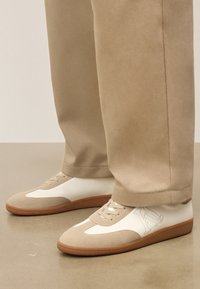 Beežid ja valged tossud, millel on kummised tallad, kantud kokku sobivate beežide pükstega siledal põrandal.