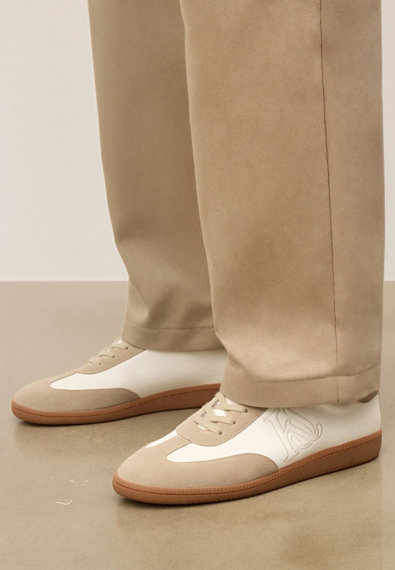 Beežid ja valged tossud, millel on kummised tallad, kantud kokku sobivate beežide pükstega siledal põrandal.