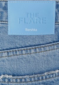 Blå denimtyg med varierande texturer och en nött detalj. En ljusblå etikett med texten "THE FLARE" och "Bershka" i präglad stil.