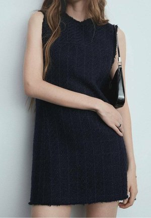 Femme portant une robe noire texturée sans manches avec un petit sac à main noir sur l'épaule et des bagues aux doigts.