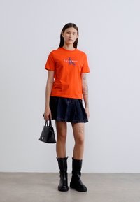 Πορτοκαλί βαμβακερό μπλουζάκι με μπλε λογότυπο, συνδυασμένο με σκούρα τζιν φούστα σε γραμμή Α και μαύρες δερμάτινες μπότες μέχρι το γόνατο, κρατώντας μία μικρή μαύρη τσάντα.