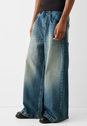 Wide-leg denim jeans in a light to dark blue gradient, featuring large side pockets and a heavy texture. Paired with black footwear.