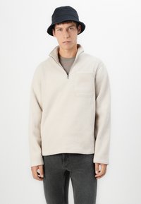 Polarowy sweter w jasnym beżu z ćwierć zamkiem, kieszenią z przodu i teksturowanym materiałem. Połączony z ciemnymi dżinsami i czarnym kapeluszem typu bucket.