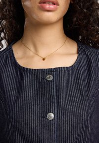 Tummansininen mekko, jossa on pystysuorat raidat, pyöreä pääntie ja hopeiset napit. Vieno kultaketju, jossa on pieni riipus, on käytössä.