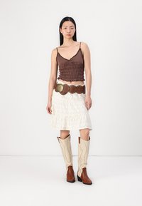 Haut smocké marron, jupe blanche en broderie anglaise avec volants, bottes de cowboy marron et beige avec des coutures décoratives. Les accessoires comprennent une ceinture marron.