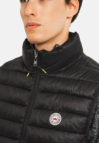 Veste matelassée noire avec col montant et fermeture zippée. Présente des cordons de serrage jaunes et un patch logo rond sur la poitrine. Intérieur gris texturé.