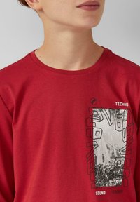 Rode longsleeve t-shirt met ronde hals, voorzien van een zwart grafisch ontwerp dat geluidsgolven en geometrische vormen op de voorkant afbeeldt.