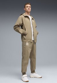 Μπεζ μπουφάν με κουκούλα και αντίστοιχο παντελόνι από ελαφρύ ύφασμα. Το σύνολο διαθέτει ελαστικές μανσέτες και λευκό λογότυπο στο παντελόνι. Άσπρα αθλητικά παπούτσια.