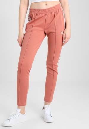 Vrouw draagt koraalroze Adidas trainingsbroek met witte strepen en logo, gecombineerd met witte sneakers en een cropped wit topje.