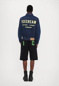 Denimjakke med overdimensjonert gul tekst "ISCREAM FOTFEST KLÆR SKATEBORD" på baksiden, sammen med svarte shorts med grønn skrift.