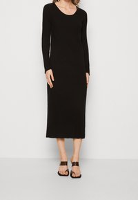 Robe noire à manches longues côtelée avec un décolleté en U et un ourlet festonné, réalisée en tissu extensible. Portée avec des sandales plates noires.