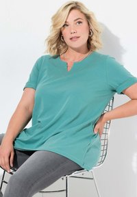 T-shirt teal a maniche corte con scollatura a V, realizzato in tessuto morbido e caratterizzato da una vestibilità comoda. Abbinato a jeans grigi, seduto su una sedia di filo.