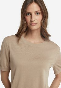Beige kortærmet striktrøje med ribbet rund hals. Glat tekstur og tætsiddende pasform, med diskrete syninger nær ærmerne.