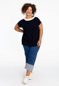 Marineblaues T-Shirt mit weißem Halsausschnitt, lockerer Schnitt, kombiniert mit verkürzten Jeans und umgeschlagenem Saum sowie weißen Sneakern. Glatte Textur.