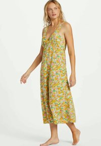 Mujer caminando descalza, vestida con un vestido midi sin mangas de color amarillo con un patrón floral en tonos rosa y blanco, frente a un fondo liso.