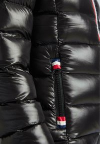 Detailní záběr na černou lesklou prošívanou bundu s vertikálním zipem zdobeným červenými, bílými a modrými textilními poutky.