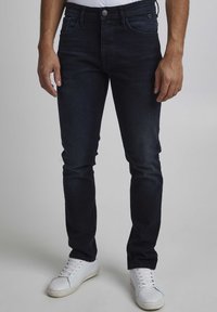 Jeans de mezclilla azul oscuro con corte ajustado, bolsillos frontales, cierre de botón plateado y un sutil detalle de desgaste, combinados con zapatillas blancas.