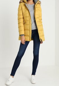 Gelbe Steppjacke mit Kapuze und Kunstfellbesatz, gestepptes Design, getragen über einem grauen Pullover, kombiniert mit dunklen Jeans und weißen Sneakers.