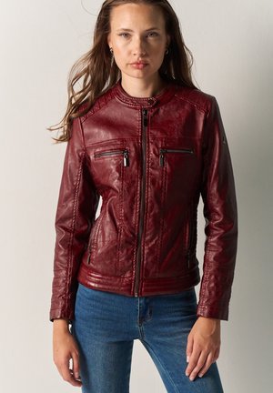 Femme portant une veste en cuir rouge foncé cintrée avec quatre poches zippées et un jean bleu, debout devant un fond uni.
