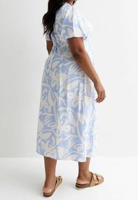 Sininen kukallinen midi-mekko lyhyillä hihoilla, jossa on löysä malli ja solmitys yksityiskohtana takana. Yhdistetty ruskeisiin liukuvaatimiin.