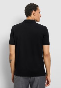 Man die van achteren gezien is, draagt een zwarte polo met korte mouwen en grijze broek, staand tegen een eenvoudige lichtgrijze achtergrond.