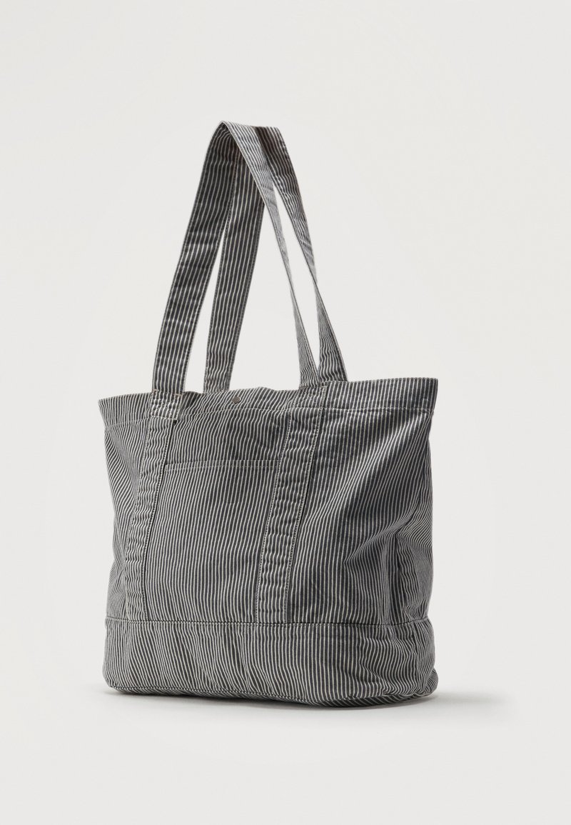 Sac fourre-tout en tissu rayé vertical noir et blanc avec deux longues anses et une poche frontale, debout sur un fond uni.