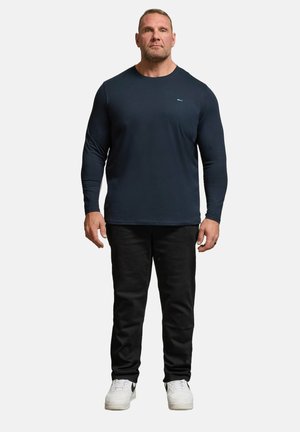 Homme debout de face, portant une chemise bleu marine ajustée à manches longues, un pantalon noir et des baskets blanches sur un fond clair uni.