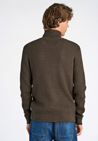 Brauner Strickpullover mit strukturiertem Rautenmuster, hohem Kragen und gerippten Bündchen und Saum, kombiniert mit blauen Jeans.
