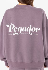 Lilla hettegenser med avslappet passform, ribbede mansjetter og bakgrunnsgrafikk med teksten "Pegador" og tilleggssitater under i hvitt.