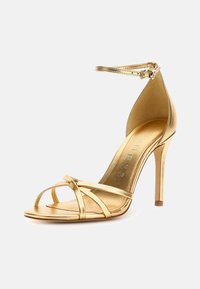 Sandal in metallo oro con tacco alto, fasce sottili, punta aperta, cinturino alla caviglia con fibbia e design affusolato. Finitura interna morbida.