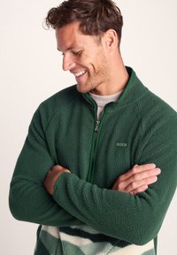 Green fleece jacket with a zippered front, textured fabric, and ribbed cuffs. Features a subtle logo patch on the chest.
