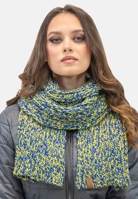 Gestrickter Schal in Blau und Gelb, mit einem strukturierten Muster und einem Lederetikett am Ende. Wird über einer grauen Daunenjacke getragen.