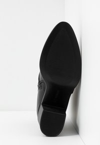 Zapato de piel negra con punta afilada, que presenta una suela negra texturizada y un tacón modesto, mostrado desde un ángulo lateral sobre un fondo blanco.