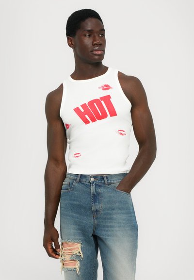 Witte tanktop met rode "HOT" opdruk en rode lippenkuspatronen. Gecombineerd met versleten blauwe jeans met gescheurde plekken op één knie.