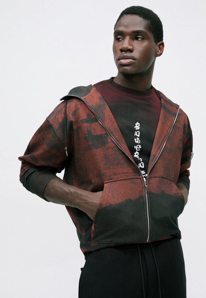 Ung mand med hænderne i lommerne iført en rød og sort tie-dye zip hoodie over en mørk skjorte med hvid lodret tekst.